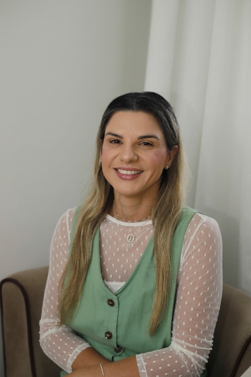 Jamille Morelli, consultora financeira, sentada em uma poltrona com blusa branca e colete verde